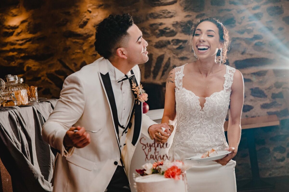 Braut und Bräutigam lachen fröhlich, während sie während ihrer standesamtlichen Trauung ihre Hochzeitstorte anschneiden. Der Bräutigam im weißen Smoking füttert die Braut, die ein Spitzenhochzeitskleid trägt, spielerisch mit Kuchen. Sie stehen vor einer strukturierten Steinmauer mit gedämpftem Licht, was eine warme und intime Atmosphäre schafft. - Elis Wedding - Hochzeitsfotograf Frankfurt