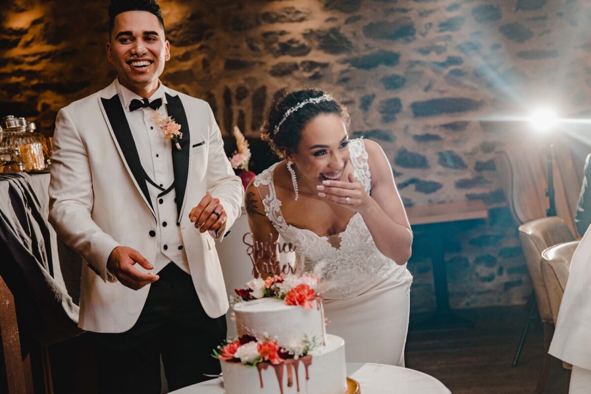 Eine Braut im weißen Spitzenkleid und ein Bräutigam im weißen Smoking mit schwarzem Revers stehen lachend hinter einer dekorierten, zweistöckigen Hochzeitstorte. Die Braut hält sich die Hand vor den Mund. Sie befinden sich während ihres Fotoshootings in einem warm beleuchteten Raum mit Steinwänden und einem Scheinwerfer, der aus dem Hintergrund leuchtet. - Elis Wedding - Hochzeitsfotograf Frankfurt