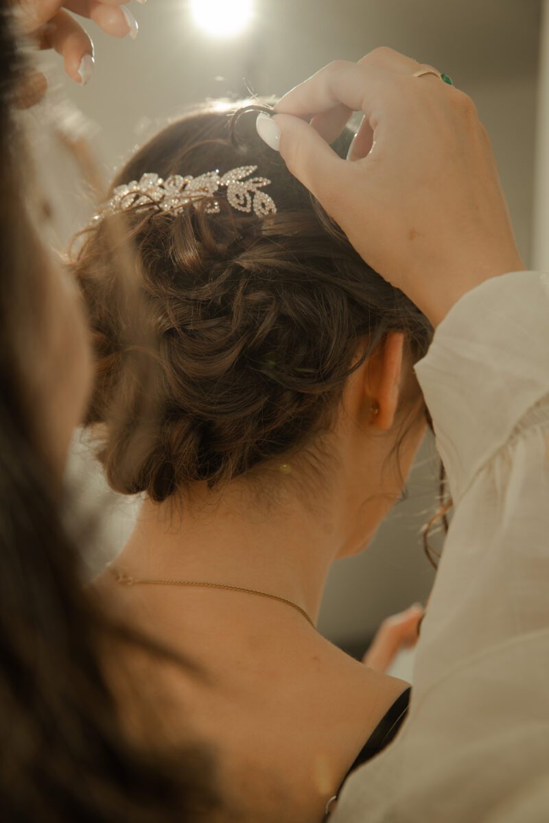 Eine Person mit dunklem Haar lässt sich während eines Fotoshootings eine elegante Hochsteckfrisur machen. Eine andere Person richtet ein dekoratives, mit Edelsteinen verziertes Haarteil mit Blumenmustern. Die Beleuchtung hebt die komplizierten Details des Haarteils hervor und verleiht der Szene einen warmen Glanz. - Elis Wedding - Hochzeitsfotograf Frankfurt