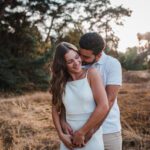 Ein Paar umarmt sich liebevoll im Freien mit trockenem Gras und Bäumen im Hintergrund. Die Frau, gekleidet in ein weißes Kleid, lächelt mit geschlossenen Augen, während der Mann im weißen Hemd hinter ihr steht, sie umarmt und ihr einen Kuss auf die Wange gibt. Dieser heitere Moment fängt die Essenz eines Outdoor-Fotoshootings in Frankfurt ein. - Elis Wedding - Hochzeitsfotograf Frankfurt