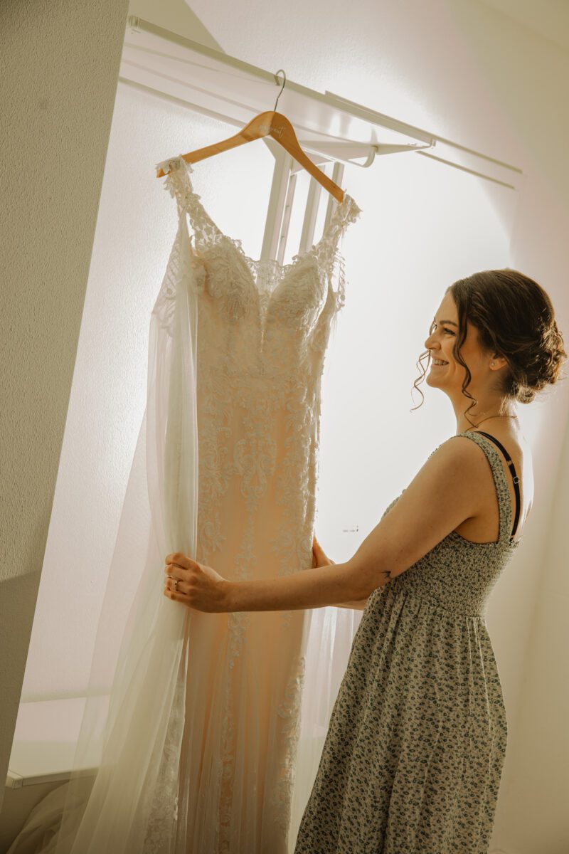 Eine Frau steht in einem sanft beleuchteten Raum und hält ein weißes Spitzenhochzeitskleid, das an einem hölzernen Kleiderbügel hängt. Sie ist im Profil zu sehen und lächelt, während sie das Kleid betrachtet. Sie hat dunkles Haar, das zu einer lockeren Hochsteckfrisur frisiert ist, und trägt ein graues ärmelloses Kleid mit Blumenmuster – ein perfekter Moment für jeden Hochzeitsfotografen. - Elis Wedding - Hochzeitsfotograf Frankfurt