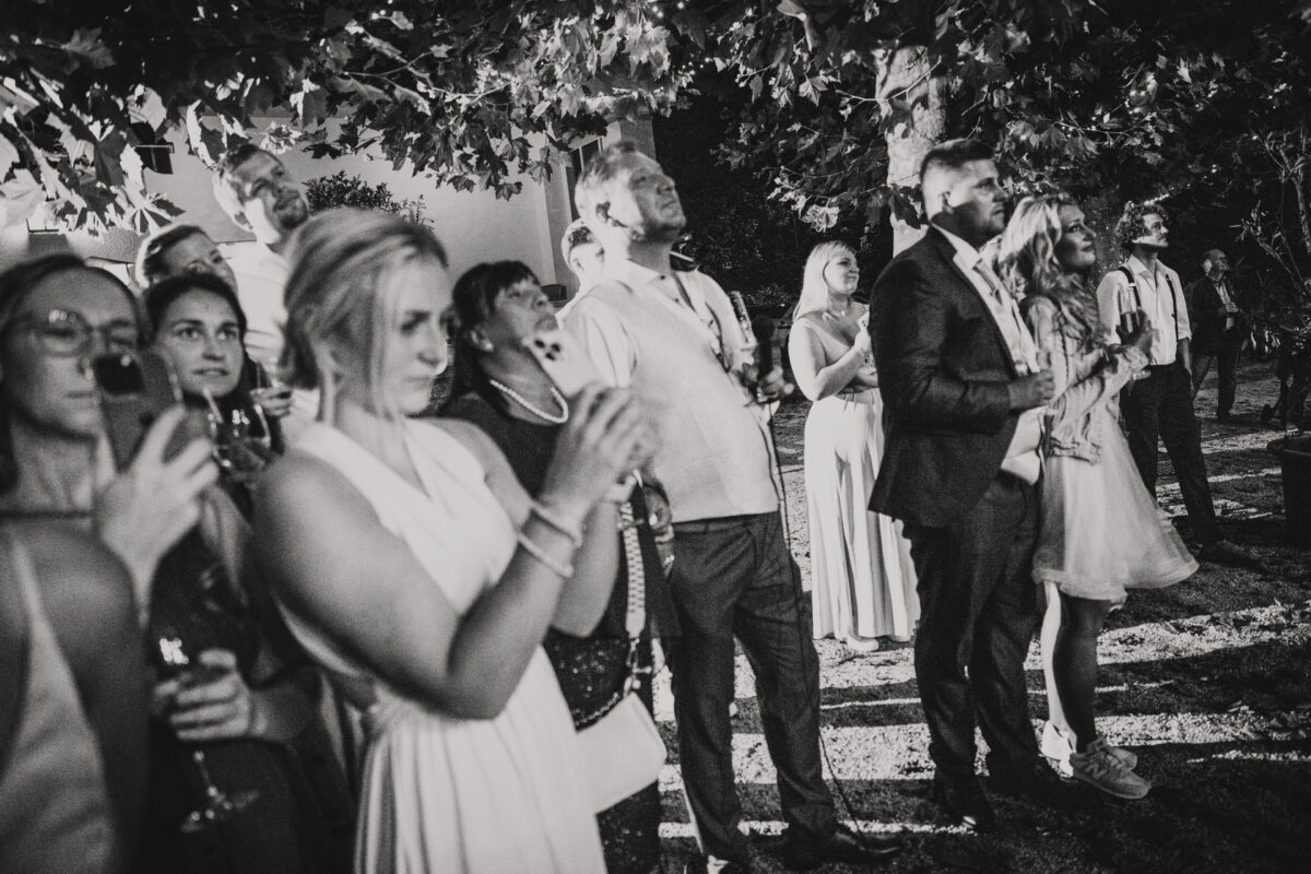Ein Schwarzweißfoto zeigt eine Gruppe von Menschen im Freien bei Nacht in Frankfurt. Die Personen stehen dicht beieinander, blicken nach oben und einige halten den Moment mit ihren Handys oder Kameras fest. Sie sind formell gekleidet, die Männer tragen Anzüge und die Frauen Kleider, was auf einen besonderen Anlass oder eine Feier schließen lässt. - Elis Wedding - Hochzeitsfotograf Frankfurt