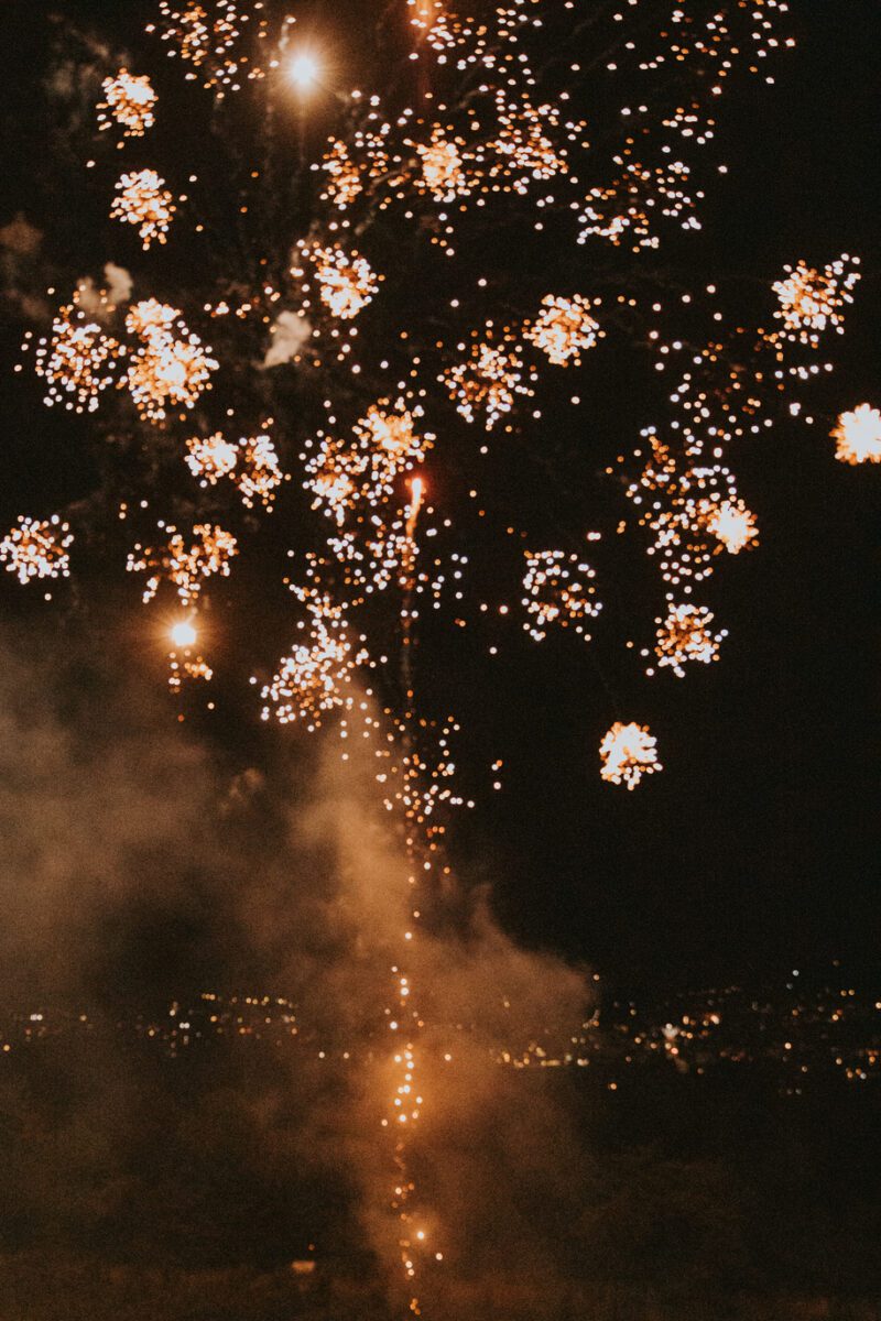 Ein Nachthimmel über Frankfurt, der von leuchtenden goldenen Feuerwerksexplosionen erhellt wird. Das Feuerwerk erzeugt funkelnde Muster, aus denen helle Lichter strahlen. Darunter sind schwache Lichter der Stadt zu sehen, und es gibt eine dunstige Rauchspur von den Feuerwerken, die der Fotografie Tiefe und Dynamik verleiht. - Elis Wedding - Hochzeitsfotograf Frankfurt