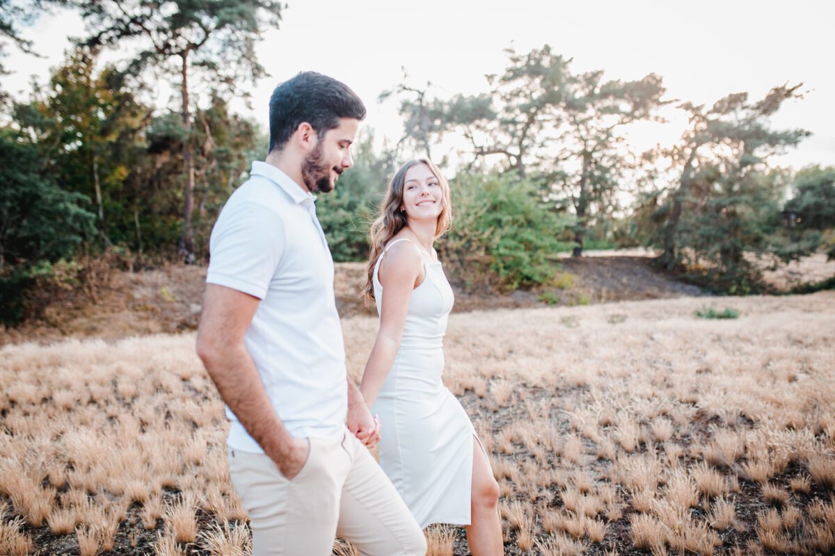 Ein Paar geht während des Fotoshootings Hand in Hand durch ein Feld mit trockenem, goldenem Gras. Beide tragen helle, legere Sommerkleidung. Die Frau lächelt strahlend, während sie in die Kamera blickt, und der Mann lächelt, während er sie ansieht. Im Hintergrund sind hohe grüne Bäume unter einem hellen Himmel zu sehen. - Elis Wedding - Hochzeitsfotograf Frankfurt