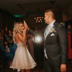 Ein frisch vermähltes Paar, eine Braut in einem kurzen weißen Kleid und ein Bräutigam in einem blauen Anzug, geben sich auf der Tanzfläche ein High-Five. Sie sind umgeben von lächelnden Gästen in Frankfurt. Der Hintergrund ist warm beleuchtet und schafft eine festliche und fröhliche Atmosphäre, perfekt für unvergessliche Fotos. - Elis Wedding - Hochzeitsfotograf Frankfurt