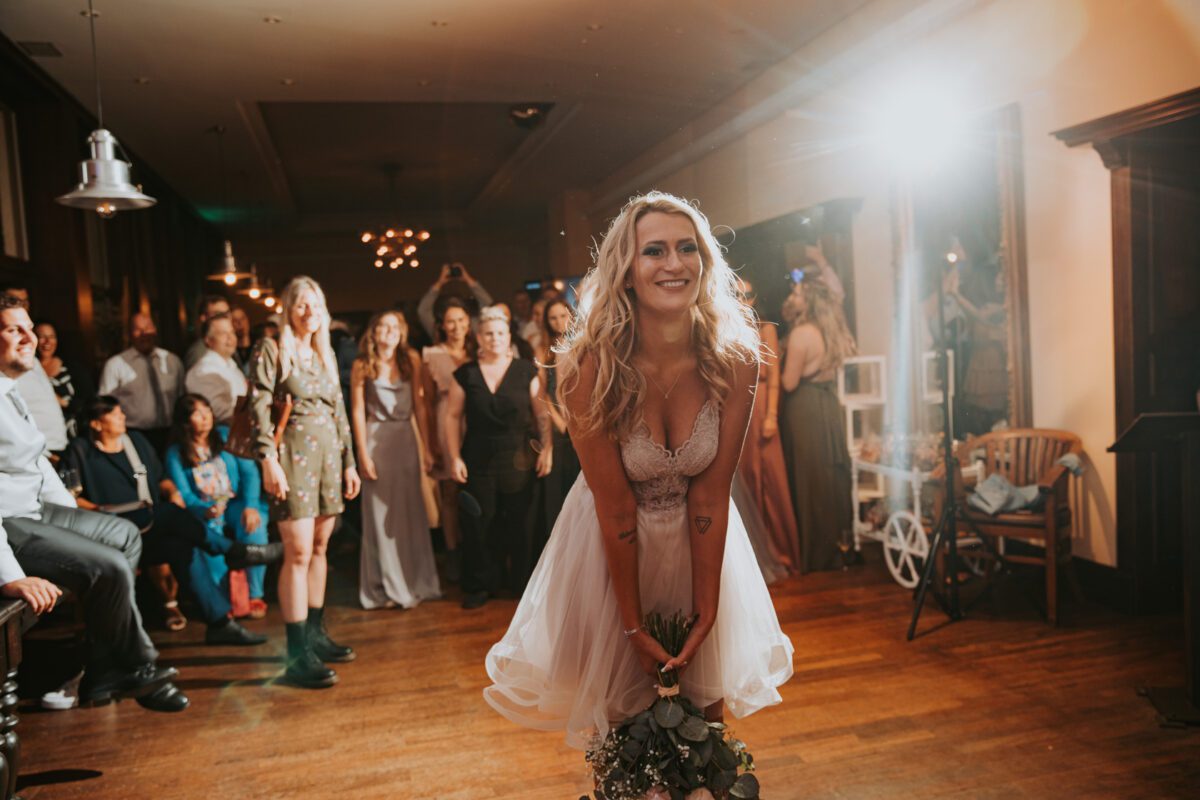 Eine lächelnde Braut in einem weißen Kleid bereitet sich darauf vor, ihren Brautstrauß in einen schwach beleuchteten Raum voller Hochzeitsgäste zu werfen. In diesem wunderschönen Moment, der durch Fotografie festgehalten wurde, steht sie im Vordergrund und beugt sich leicht vor, mit dem Brautstrauß in den Händen. Gäste, darunter leger und formell gekleidete Männer und Frauen, schauen mit eifrigen Mienen aus dem Hintergrund zu. - Elis Wedding - Hochzeitsfotograf Frankfurt