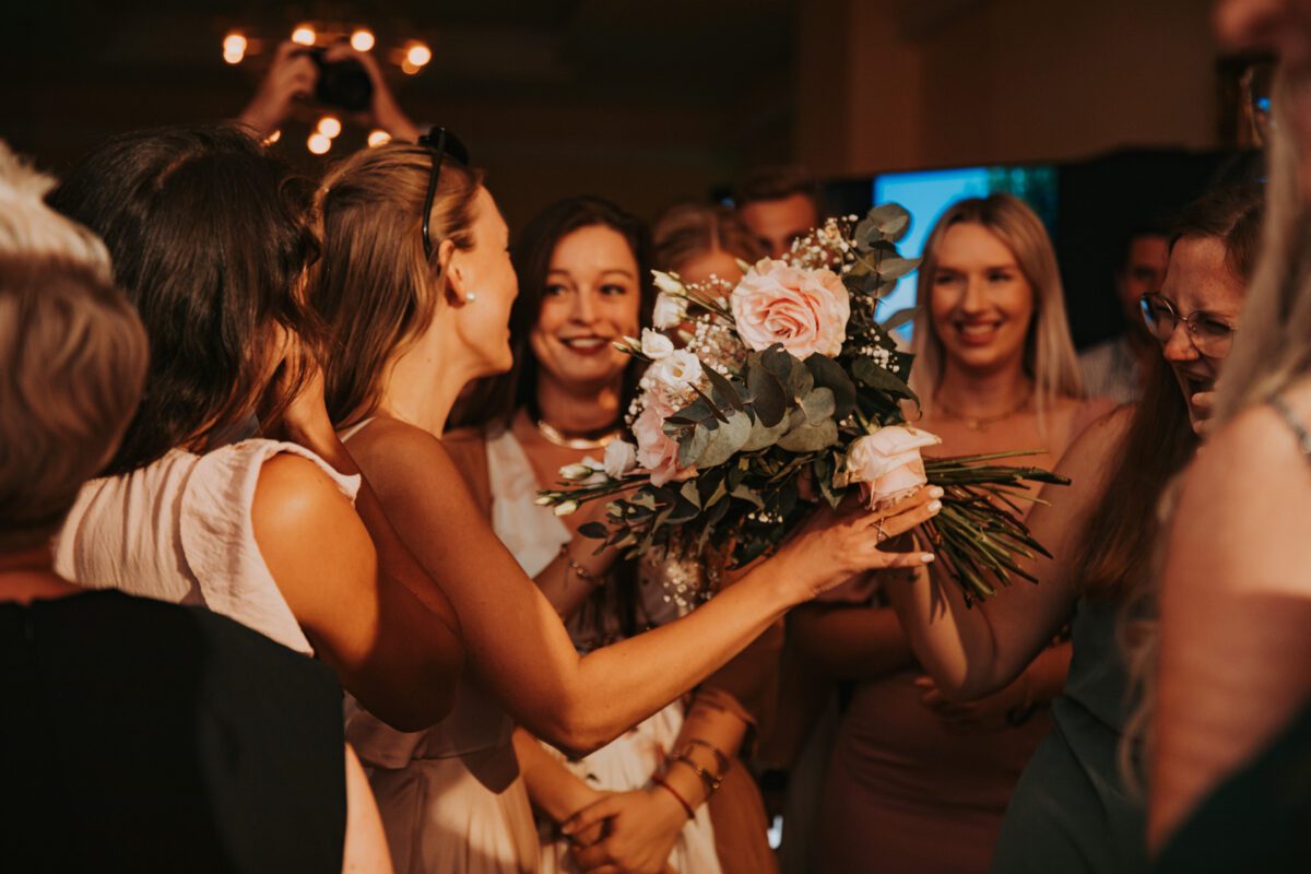 Eine Gruppe lächelnder Frauen, meist in hellen Kleidern, versammelt sich um eine Frau, die einen Strauß rosa Rosen und Eukalyptusblätter hält. Der Raum ist warm beleuchtet und unscharfe Figuren im Hintergrund lassen darauf schließen, dass es sich um eine feierliche Veranstaltung handelt, möglicherweise eine Hochzeit. Aufgenommen von einem Hochzeitsfotografen in Frankfurt, unterstreichen freudige Ausdrücke die festliche Atmosphäre. - Elis Wedding - Hochzeitsfotograf Frankfurt