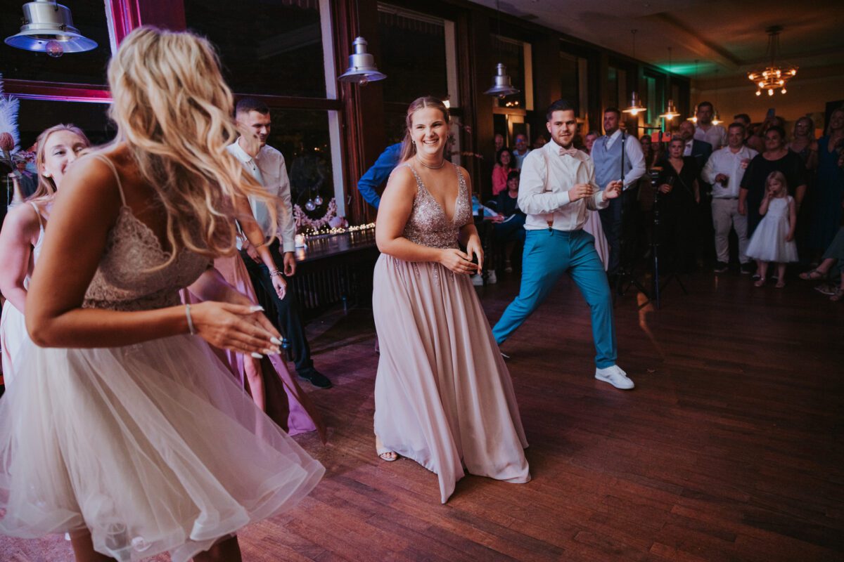 Eine lebhafte Tanzszene bei einer Hochzeit in Frankfurt. Männer und Frauen in formeller Kleidung tanzen voller Begeisterung. Zwei Frauen im Vordergrund tragen helle Kleider und ein Mann in weißem Hemd und blauer Hose tanzt neben ihnen. Gäste im Hintergrund schauen zu und lächeln, während ihr Hochzeitsfotograf den Moment festhält. Der Raum ist warm beleuchtet. - Elis Wedding - Hochzeitsfotograf Frankfurt