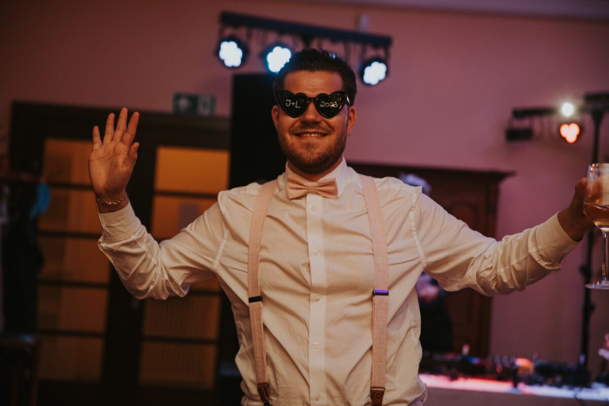 Ein Mann mit einer neuartigen Brille aus dem Jahr 2021 steht mit erhobenen Armen da und ist bereit für ein Fotoshooting. Er trägt ein weißes Oberhemd mit einer rosa Fliege und Hosenträgern und hält ein Getränk in einer Hand. Im Hintergrund sind Partylichter und verschwommene Dekorationen zu sehen, die eine festliche Feieratmosphäre im Innenbereich suggerieren. - Elis Wedding - Hochzeitsfotograf Frankfurt