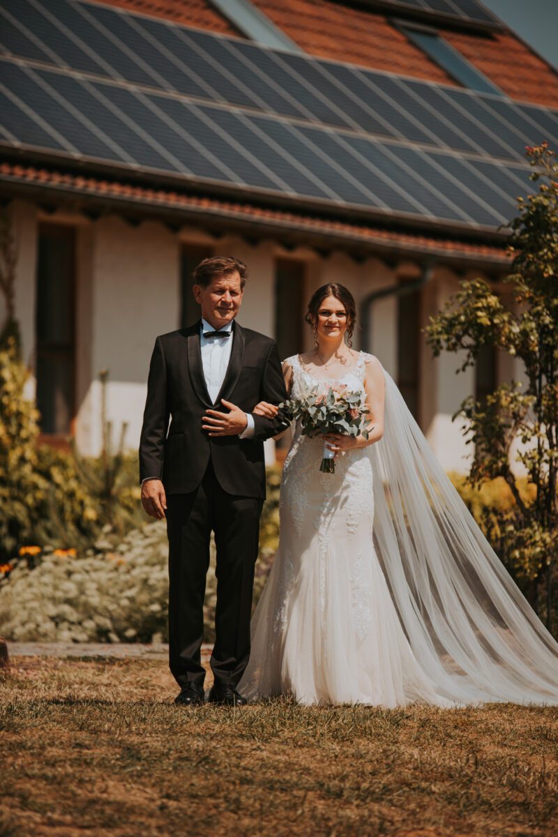 Eine Braut in einem weißen Brautkleid mit langem Schleier geht während ihrer standesamtlichen Trauung Arm in Arm mit einem älteren Mann in schwarzem Anzug und Fliege. Die Braut hält einen Blumenstrauß in der Hand, während sie über den Rasen spazieren und Momente für ihr Fotoshooting vor der Kulisse von Blumen, Pflanzen und Gebäuden mit Solarpanelen festhalten. - Elis Wedding - Hochzeitsfotograf Frankfurt