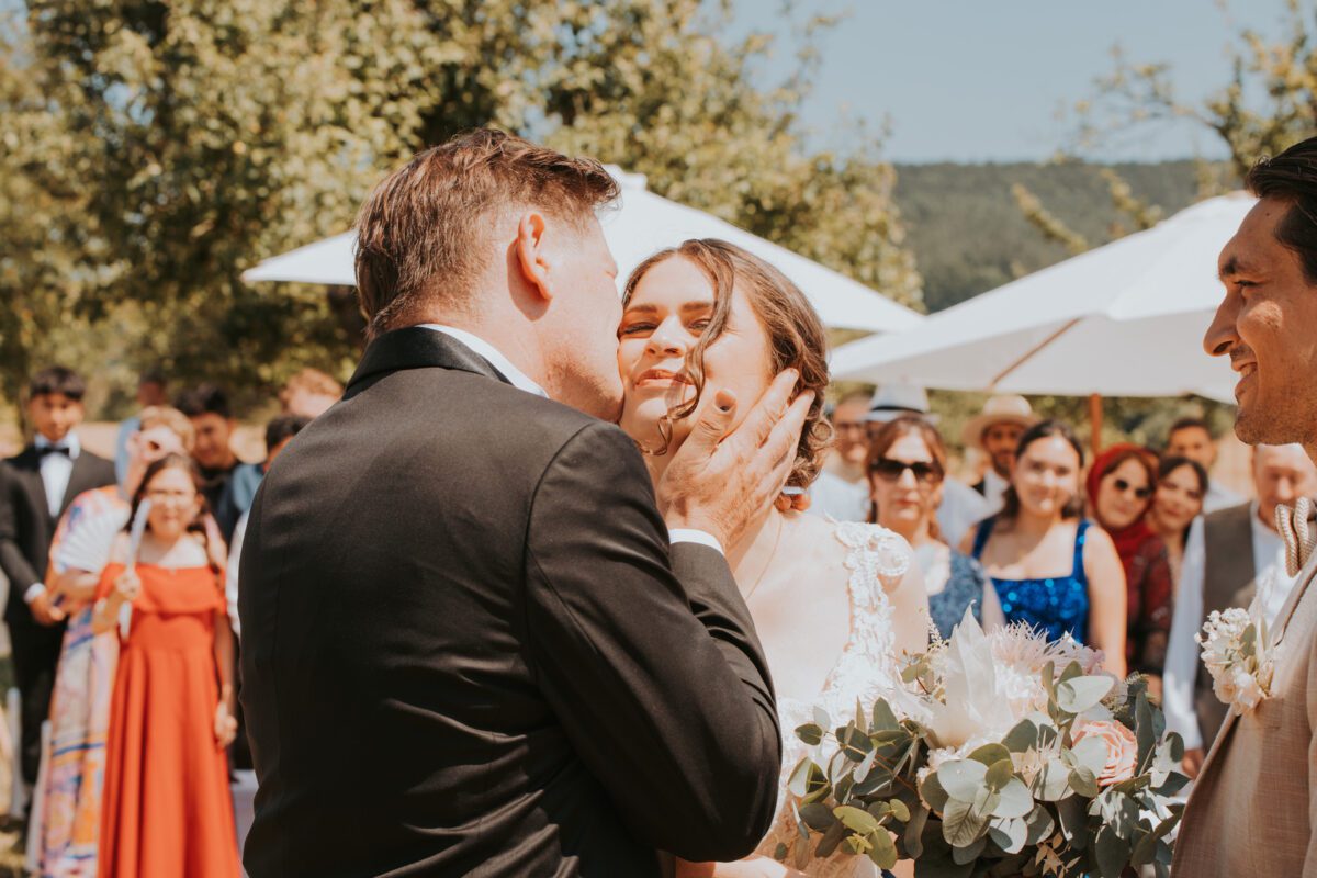 Eine Braut, die einen Blumenstrauß in der Hand hält und ein weißes Brautkleid trägt, bekommt von einem Mann im schwarzen Anzug einen zärtlichen Kuss auf die Wange. Im Hintergrund stehen Gäste, einige halten Sonnenschirme, beobachten die Szene und lächeln. Die Außenkulisse in der Nähe von Frankfurt zeigt viel Grün und einen sonnigen Himmel. - Elis Wedding - Hochzeitsfotograf Frankfurt