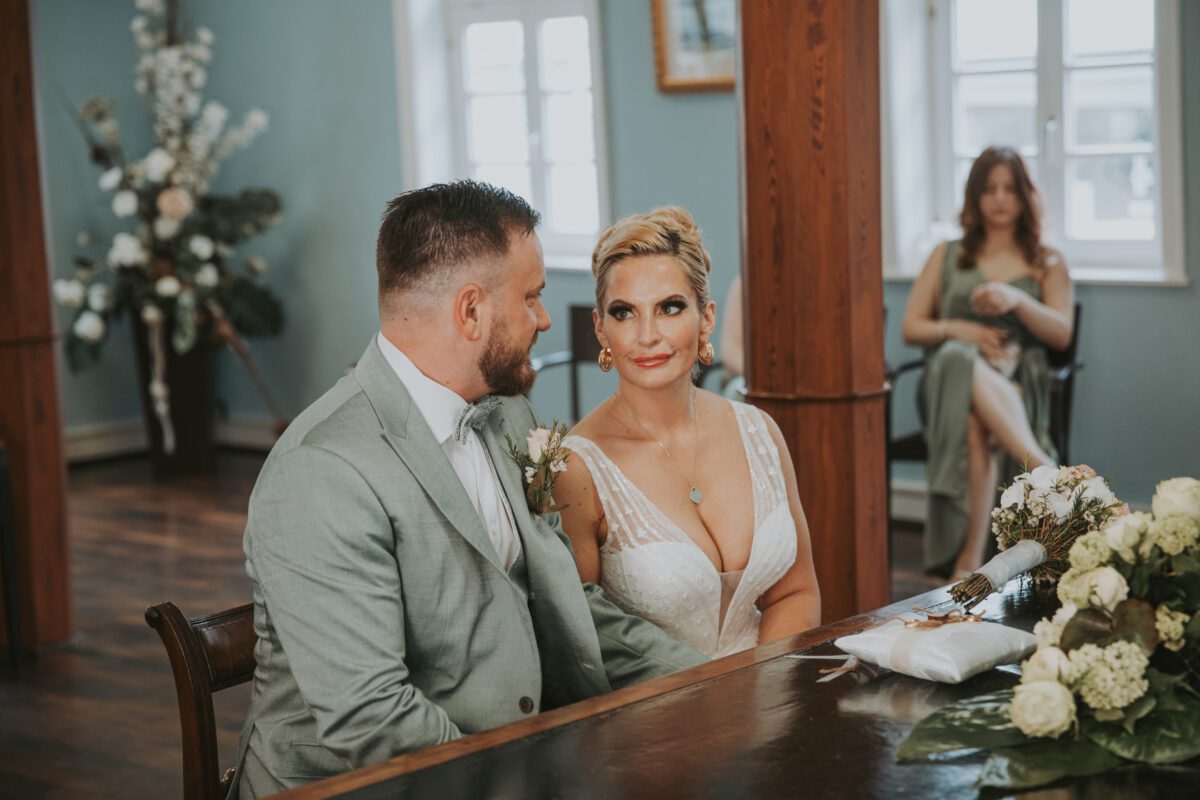 Braut und Bräutigam in Abendgarderobe sitzen während eines Fotoshootings eng aneinander gedrängt an einem mit Blumen geschmückten Holztisch. Die Braut trägt ein weißes Brautkleid mit tiefem Ausschnitt und der Bräutigam einen hellgrauen Anzug. Im Hintergrund sitzt eine Brautjungfer in einem hellgrünen Kleid auf einem Stuhl, in der Nähe des Fensters sind Blumen zu sehen. - Elis Wedding - Hochzeitsfotograf Frankfurt