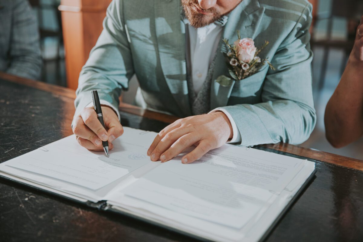 Eine Person in einem hellblauen Anzug mit einer Blumenboutonniere unterschreibt sorgfältig ein formelles Dokument auf einem schwarzen Tisch. Der Hochzeitsfotograf fängt den Moment perfekt ein, da das Gesicht der Person teilweise sichtbar ist und sie konzentriert auf das Papier fokussiert ist, während sie in ihrer rechten Hand einen Stift hält. - Elis Wedding - Hochzeitsfotograf Frankfurt