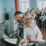 Ein Paar sitzt eng beieinander und lächelt, während einer Hochzeitszeremonie in Frankfurt hält es ein kleines Kind im Arm. Der Mann trägt einen hellgrünen Anzug und eine Fliege, die Frau ein weißes Kleid. Sie sitzen an einem Holztisch mit einem Blumenstrauß. Die Gäste hinter ihnen, in formeller Kleidung gekleidet, klatschen und lächeln in dem hellblauen Raum, während der besondere Moment fotografisch festgehalten wird. - Elis Wedding - Hochzeitsfotograf Frankfurt