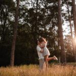 Ein Paar umarmt sich auf einer sonnenbeschienenen Waldlichtung, wunderschön eingefangen von ihrem Hochzeitsfotografen aus Frankfurt. Hohe Bäume umgeben sie, während die Sonne einen goldenen Schimmer durch die Zweige wirft. Die Frau, in einem leichten Kleid und weißen Turnschuhen, wird von dem Mann, der ein leichtes Hemd und eine leichte Hose trägt, vom Boden hochgehoben. Hohes Gras wiegt sich sanft um sie herum. - Elis Wedding - Hochzeitsfotograf Frankfurt