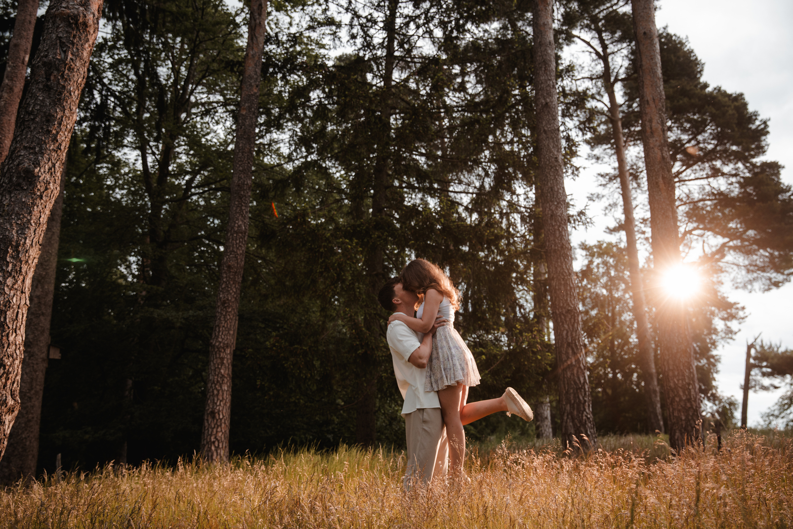 Ein Paar umarmt sich auf einer sonnenbeschienenen Waldlichtung, wunderschön eingefangen von ihrem Hochzeitsfotografen aus Frankfurt. Hohe Bäume umgeben sie, während die Sonne einen goldenen Schimmer durch die Zweige wirft. Die Frau, in einem leichten Kleid und weißen Turnschuhen, wird von dem Mann, der ein leichtes Hemd und eine leichte Hose trägt, vom Boden hochgehoben. Hohes Gras wiegt sich sanft um sie herum. - Elis Wedding - Hochzeitsfotograf Frankfurt