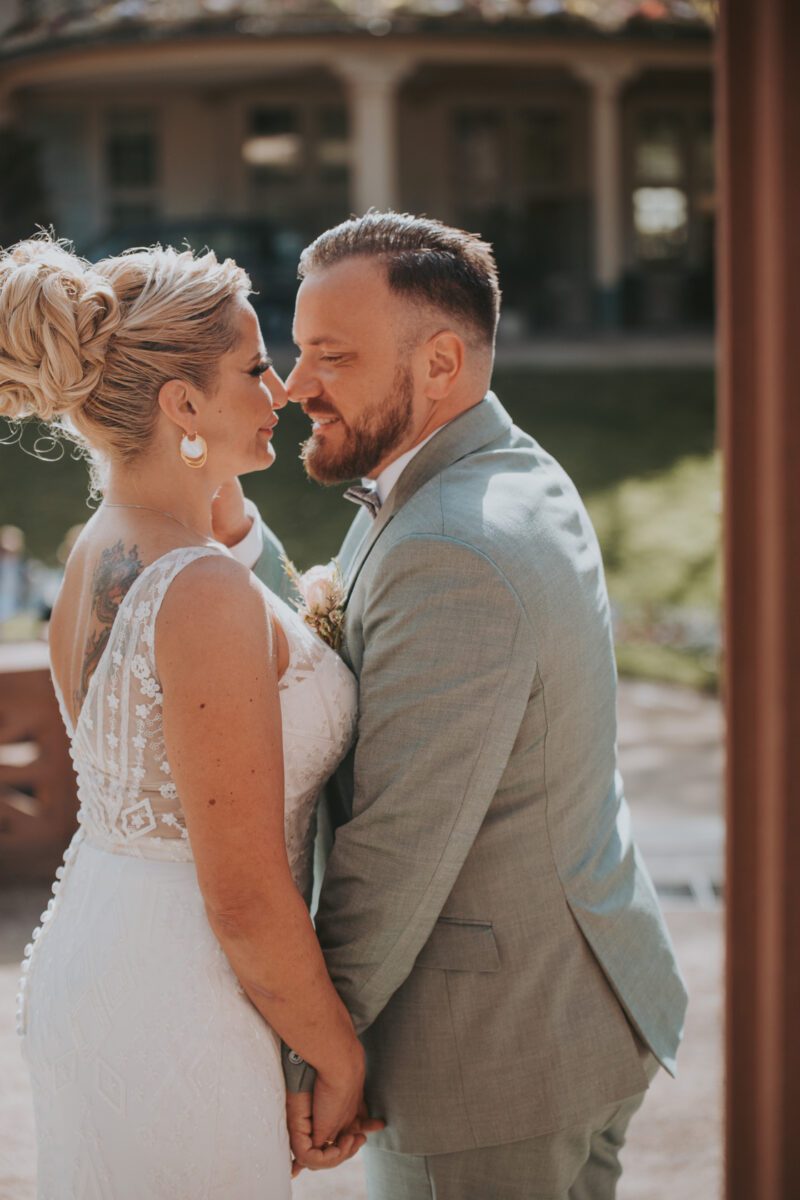 Braut und Bräutigam blicken sich während ihres Fotoshootings im Freien in Frankfurt liebevoll an. Die Braut trägt ein weißes Spitzenkleid und ihr Haar ist elegant hochgesteckt. Der Bräutigam trägt einen hellgrauen Anzug mit Fliege. Sie halten Händchen, während die Sonne einen warmen Schein auf ihre lächelnden Gesichter wirft, mit verschwommenem Grün und einem Gebäude im Hintergrund. - Elis Wedding - Hochzeitsfotograf Frankfurt