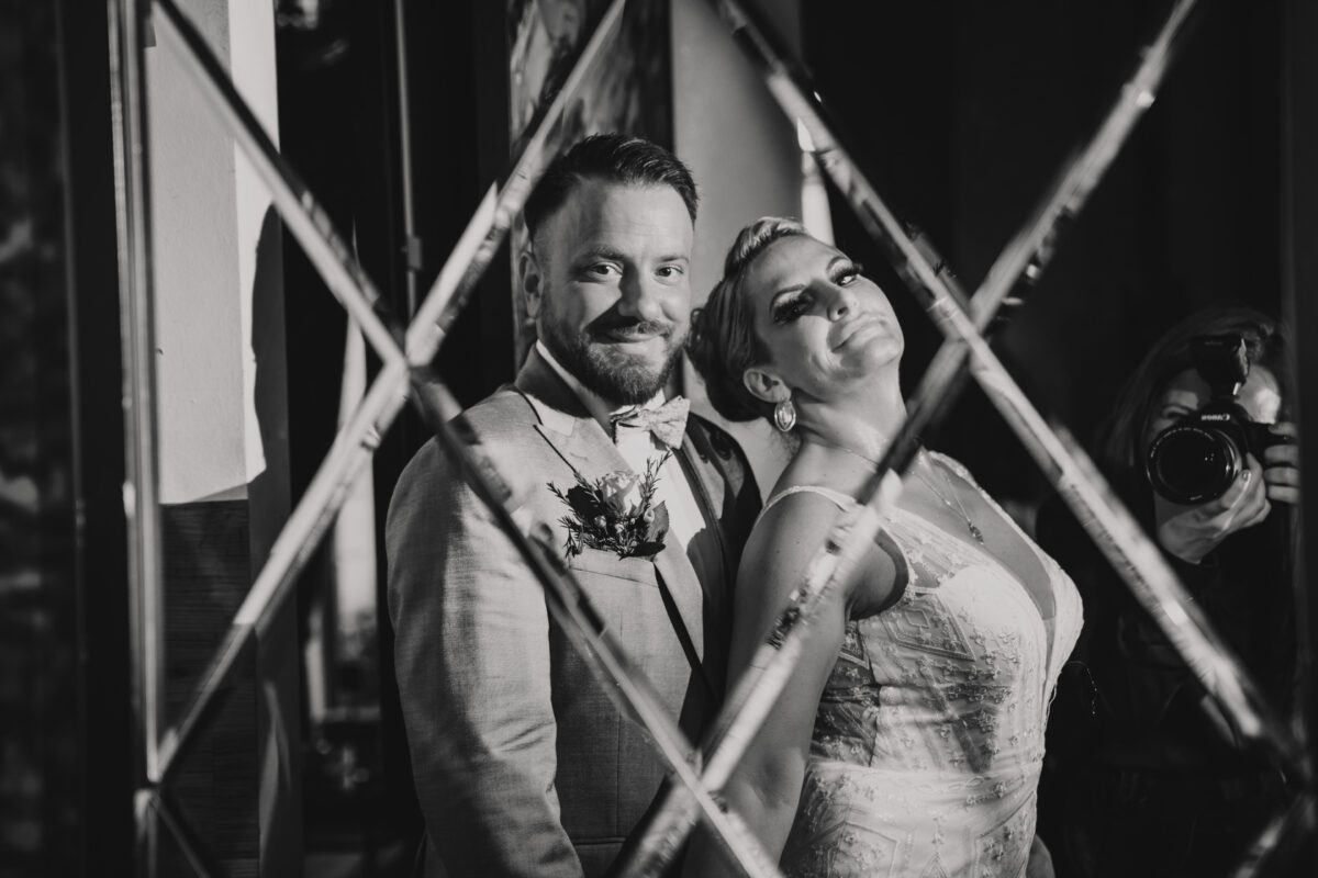Schwarzweißfoto eines lächelnden Paares an seinem Hochzeitstag. Der Bräutigam, in einem hellen Anzug mit Fliege und Ansteckblume, steht dicht neben der Braut, die ein Spitzenkleid trägt. Während die Braut sich zurücklehnt und in die Kamera lächelt, fängt diese bezaubernde Aufnahme ihres Hochzeitsfotografen die Liebe in Frankfurt wunderschön ein. - Elis Wedding - Hochzeitsfotograf Frankfurt