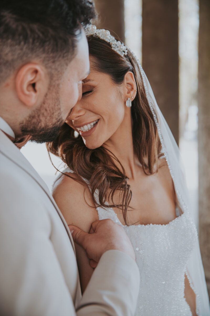 Braut und Bräutigam erleben einen intimen Moment während ihres Fotoshootings am Hochzeitstag. Die Braut, die ein funkelndes weißes Kleid und einen perlenbesetzten Kopfschmuck trägt, lächelt mit geschlossenen Augen. Der Bräutigam, der einen hellen Anzug trägt, berührt sanft ihren Arm. Beide stehen nah beieinander und strahlen Freude aus. - Elis Wedding - Hochzeitsfotograf Frankfurt