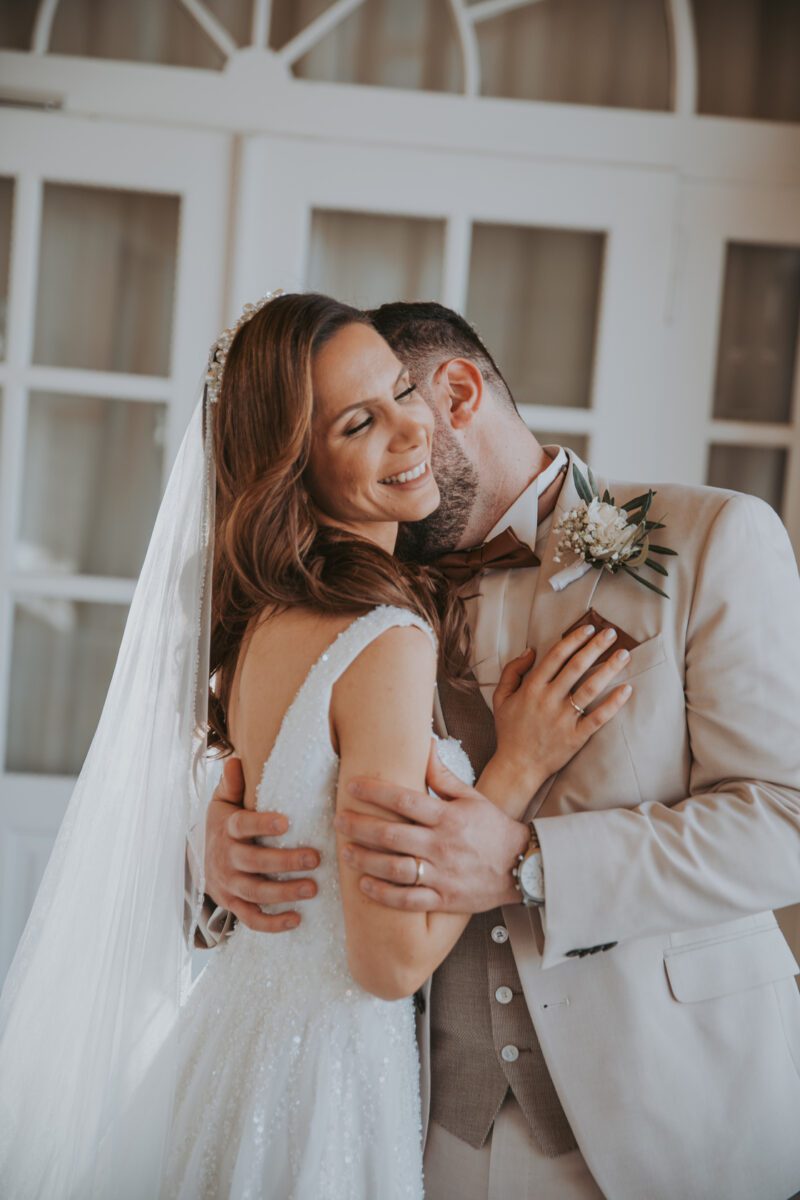 Braut und Bräutigam, die sich in Frankfurt kennengelernt haben, umarmen sich und lächeln freudig an ihrem Hochzeitstag. Die Braut, in einem weißen Kleid und Schleier, hält den Bräutigam fest. Der Bräutigam, in einem beigen Anzug mit einer braunen Fliege, hat seine Arme um sie geschlungen. Sie stehen vor weißen Fenstertüren, getaucht in sanftes Tageslicht. - Elis Wedding - Hochzeitsfotograf Frankfurt