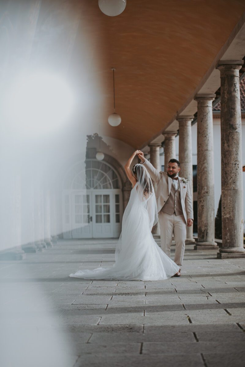 Braut und Bräutigam tanzen in einem eleganten Säulengang. Die Braut trägt ein fließendes weißes Kleid und einen Schleier, und der Bräutigam trägt einen hellen Anzug. Umgeben von hohen Marmorsäulen und einem gewölbten Eingang ist die Szene, die ein Hochzeitsfotograf aus Frankfurt eingefangen hat, sanft beleuchtet und ätherisch. - Elis Wedding - Hochzeitsfotograf Frankfurt