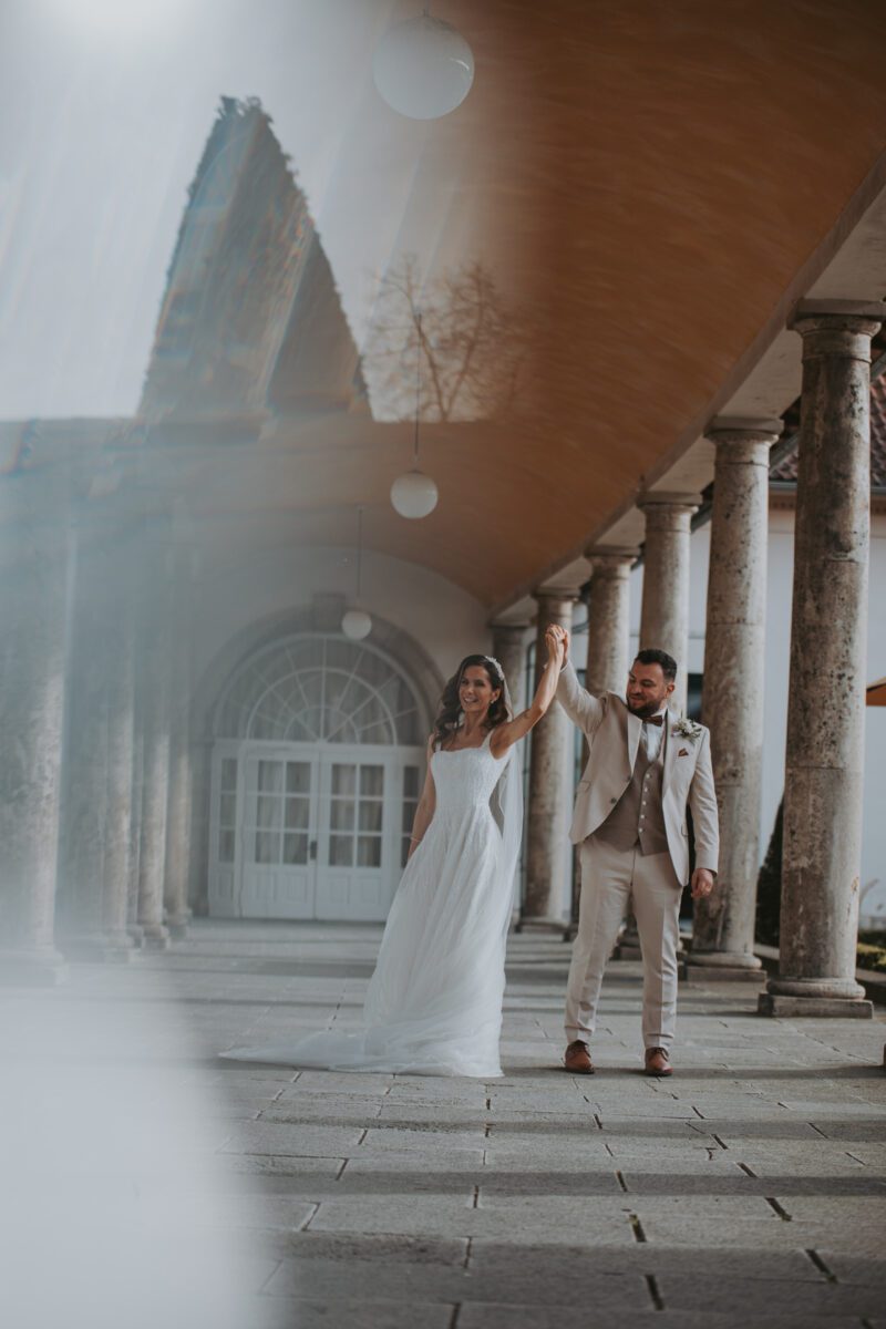 Eine Braut in einem fließenden weißen Kleid und ein Bräutigam in einem beigen Anzug halten freudig die Hände und heben sie hoch, während sie durch einen eleganten Säulengang schreiten. Zu den architektonischen Details gehören Steinsäulen und eine große gewölbte Glastür im Hintergrund. Der Hochzeitsfotograf fängt das Licht ein, das ihr Glück hervorhebt. - Elis Wedding - Hochzeitsfotograf Frankfurt