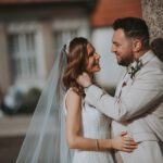 Ein Paar in Hochzeitskleidung steht dicht beieinander im Freien. Die Braut, die ein weißes Kleid und einen Schleier trägt, blickt den Bräutigam, der einen hellen Anzug trägt, liebevoll an. Sie werden während ihrer standesamtlichen Trauung von Baumstämmen eingerahmt, im Hintergrund ist ein verschwommenes Gebäude mit einem roten Ziegeldach zu sehen. - Elis Wedding - Hochzeitsfotograf Frankfurt