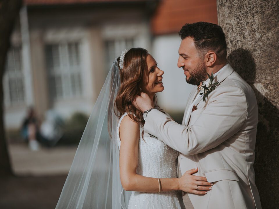 Ein Paar in Hochzeitskleidung steht dicht beieinander im Freien. Die Braut, die ein weißes Kleid und einen Schleier trägt, blickt den Bräutigam, der einen hellen Anzug trägt, liebevoll an. Sie werden während ihrer standesamtlichen Trauung von Baumstämmen eingerahmt, im Hintergrund ist ein verschwommenes Gebäude mit einem roten Ziegeldach zu sehen. - Elis Wedding - Hochzeitsfotograf Frankfurt