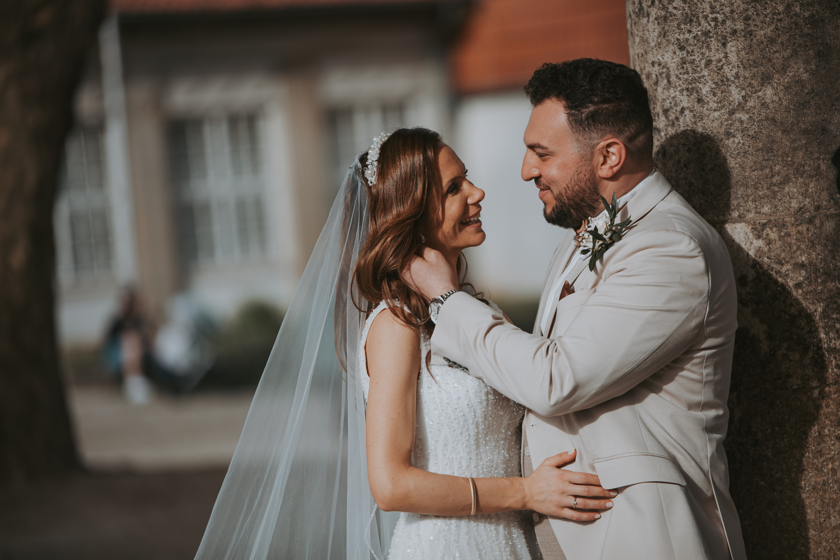 Ein Paar in Hochzeitskleidung steht dicht beieinander im Freien. Die Braut, die ein weißes Kleid und einen Schleier trägt, blickt den Bräutigam, der einen hellen Anzug trägt, liebevoll an. Sie werden während ihrer standesamtlichen Trauung von Baumstämmen eingerahmt, im Hintergrund ist ein verschwommenes Gebäude mit einem roten Ziegeldach zu sehen. - Elis Wedding - Hochzeitsfotograf Frankfurt