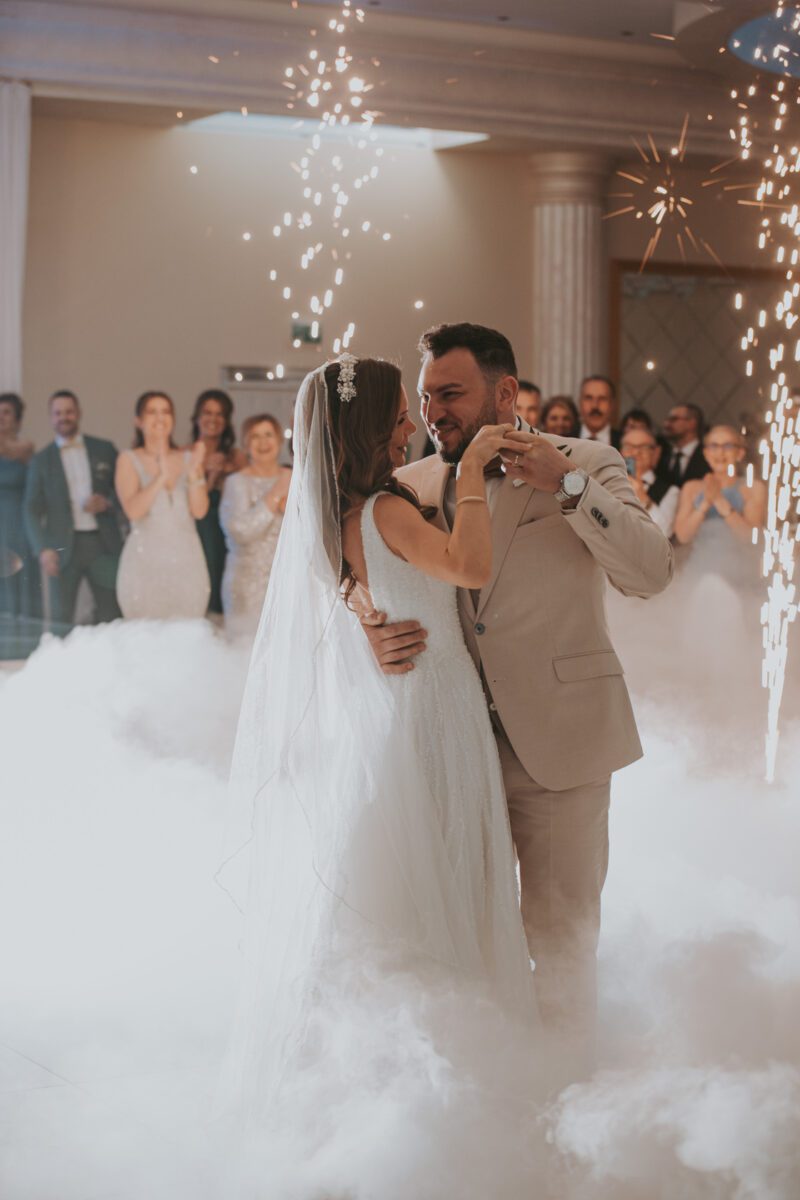 Braut und Bräutigam tanzen ihren ersten Tanz bei ihrer Hochzeit in Frankfurt, umgeben von Nebel und Pyrotechnik-Funken. Die Braut trägt ein langes, weißes Kleid und einen Schleier, während der Bräutigam einen beigen Anzug trägt. Im Hintergrund applaudieren Gäste in Abendgarderobe und beobachten das Paar mit freudigen Gesichtern. - Elis Wedding - Hochzeitsfotograf Frankfurt