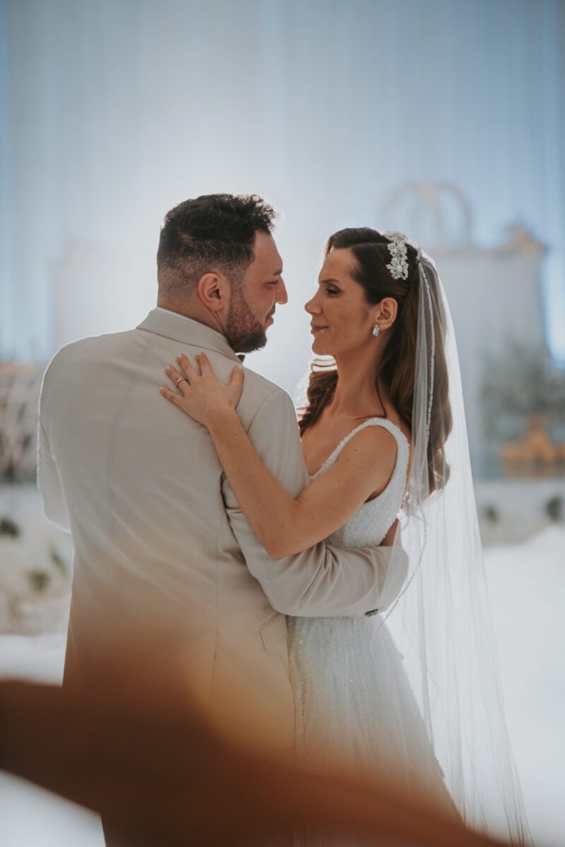 Braut und Bräutigam tanzen bei ihrer Hochzeit den ersten Tanz. Die Braut, die ein weißes Kleid und einen Schleier mit floralen Accessoires trägt, und der Bräutigam, der einen hellgrauen Anzug trägt, blicken einander in die Augen. Der Hintergrund ist sanft beleuchtet und elegant in neutralen Tönen dekoriert, perfekt für den Hochzeitsfotografen, um diesen romantischen Moment festzuhalten. - Elis Wedding - Hochzeitsfotograf Frankfurt