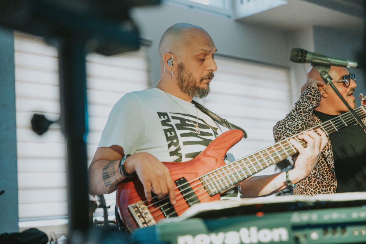 Ein kahlköpfiger Mann mit Bart spielt auf der Bühne eine E-Bassgitarre. Er trägt ein weißes Hemd und einen In-Ear-Monitor. Eine weitere Person mit Sonnenbrille und Hemd mit Leopardenmuster steht neben ihm und singt in ein Mikrofon. Im Vordergrund sind Musikgeräte und Ständer zu sehen, die die Essenz einer Live-Performance einfangen. Fotografie. - Elis Wedding - Hochzeitsfotograf Frankfurt