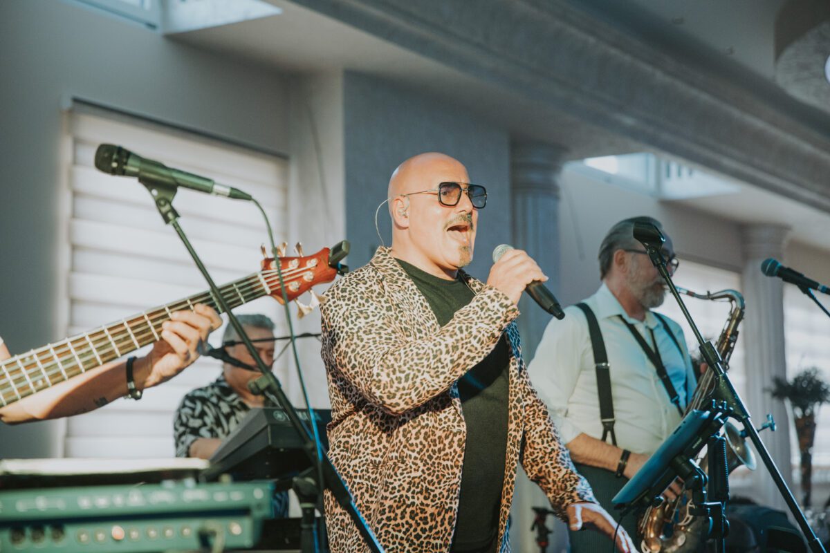 Ein kahlköpfiger Mann mit Sonnenbrille und Leopardenjacke singt leidenschaftlich in ein Mikrofon auf der Bühne eines Frankfurter Clubs. Ihm folgen links ein Bassist und rechts ein Saxophonist. Der Raum ist gut beleuchtet, Tageslicht fällt durch Jalousien. Im Hintergrund sind Musikgeräte zu sehen. - Elis Wedding - Hochzeitsfotograf Frankfurt