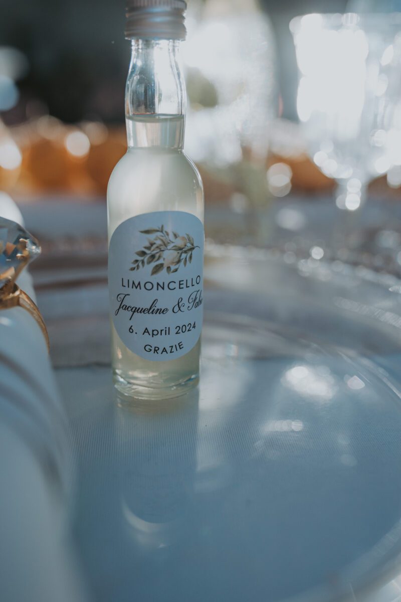 Eine kleine Flasche Limoncello mit einem personalisierten Etikett mit der Aufschrift „Limoncello, Jacqueline & John, 6. April 2024, Grazie“ steht auf einem Tisch. Die klare Flasche mit der hellgelben Flüssigkeit wurde von einem talentierten Hochzeitsfotografen aus Frankfurt aufgenommen und hebt sich von der verschwommenen Bankettkulisse mit elegantem Glasgeschirr und Dekor ab. - Elis Wedding - Hochzeitsfotograf Frankfurt