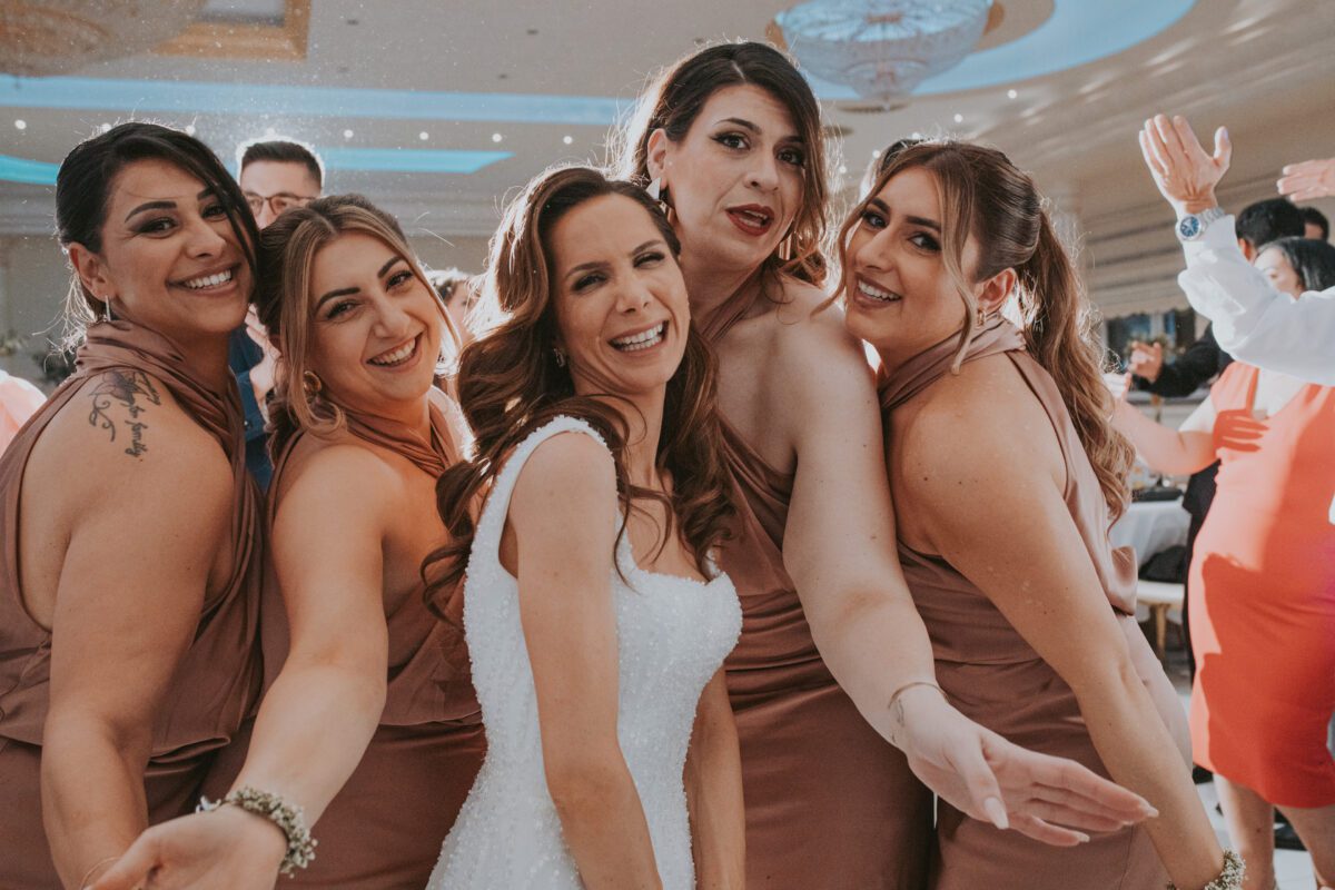 Eine fröhliche Braut in einem weißen Kleid ist bei einer Hochzeitsfeier von vier Brautjungfern in passenden lilafarbenen Kleidern umgeben. Sie posieren eng beieinander und lächeln während ihres Fotoshootings in Frankfurt in die Kamera. Im Hintergrund sieht man andere Gäste, die unter warmem, stimmungsvollem Licht feiern. - Elis Wedding - Hochzeitsfotograf Frankfurt