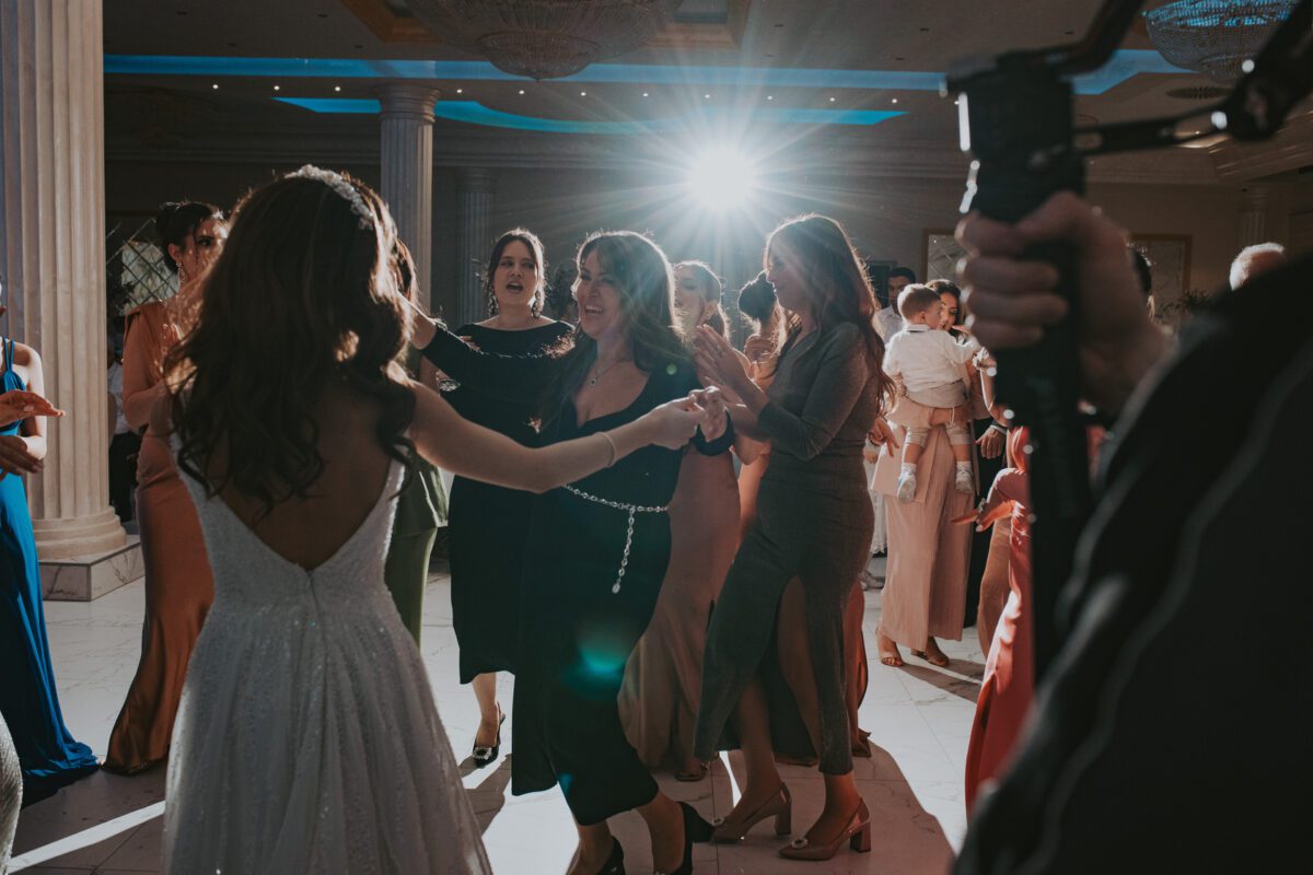 Eine Gruppe von Menschen, hauptsächlich Frauen, tanzt fröhlich zusammen bei einer Indoor-Veranstaltung. Die zentrale Figur, die ein tiefblaues Kleid trägt, hält die Hand einer Frau in einem weißen, rückenfreien Kleid. Die Stimmung ist festlich mit warmem Licht und verzierten Säulen im Hintergrund. Fotografie fängt das helle Licht ein, das hinter ihnen scheint. - Elis Wedding - Hochzeitsfotograf Frankfurt