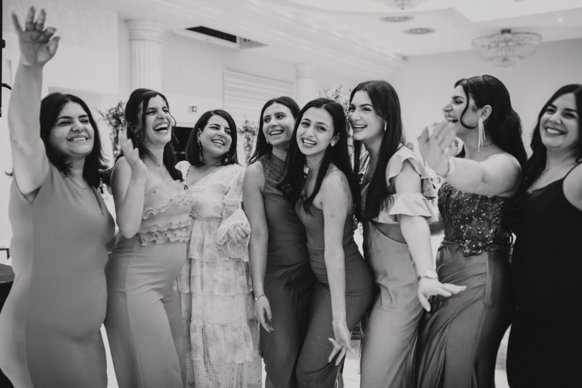 Eine Gruppe von neun Frauen steht dicht beieinander und lächelt und lacht in einer feierlichen Pose. Sie sind in elegante Abendkleider gekleidet, viele tragen lange, fließende Kleider. Die Kulisse scheint drinnen zu sein, mit kunstvollem Dekor, darunter Kronleuchter und Säulen. Die Stimmung ist festlich und fröhlich, perfekt für einen Hochzeitsfotografen. - Elis Wedding - Hochzeitsfotograf Frankfurt