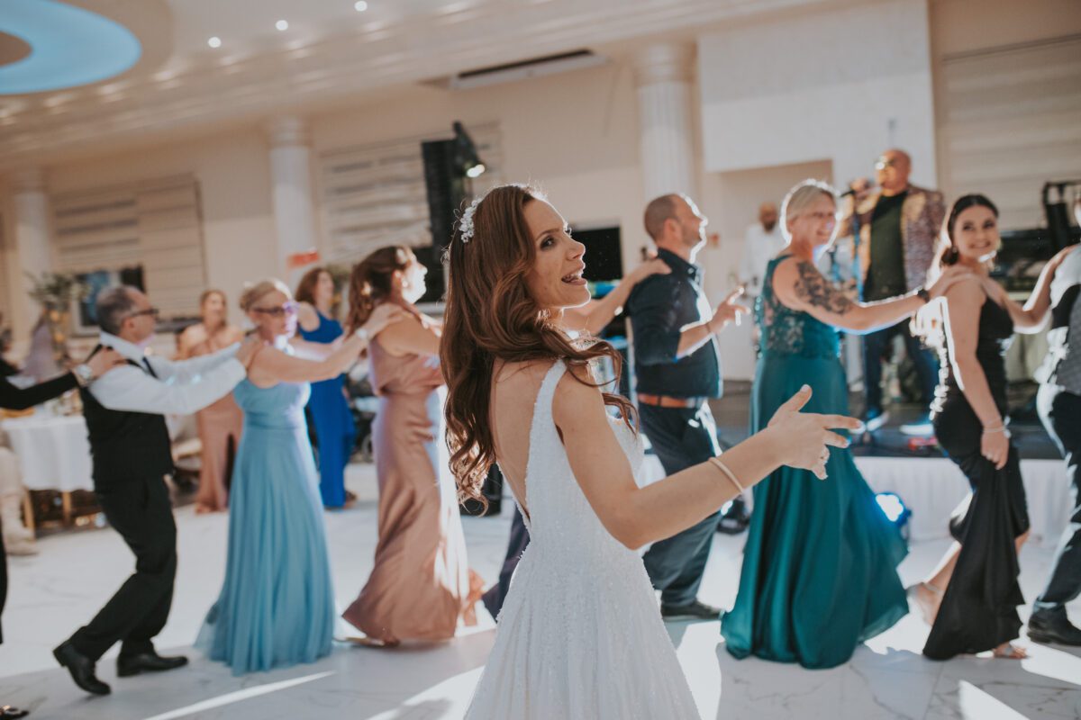 Eine fröhliche Braut in einem weißen Kleid führt einen Line Dance auf der Hochzeitsfeier an, lächelt und gestikuliert mit ihren Händen. Hinter ihr mischen sich Gäste in eleganten Kleidern und Anzügen in das Vergnügen ein. Der Raum, perfekt vom Hochzeitsfotografen eingefangen, zeichnet sich durch helles Licht, Säulen und eine Live-Band im Hintergrund aus. - Elis Wedding - Hochzeitsfotograf Frankfurt
