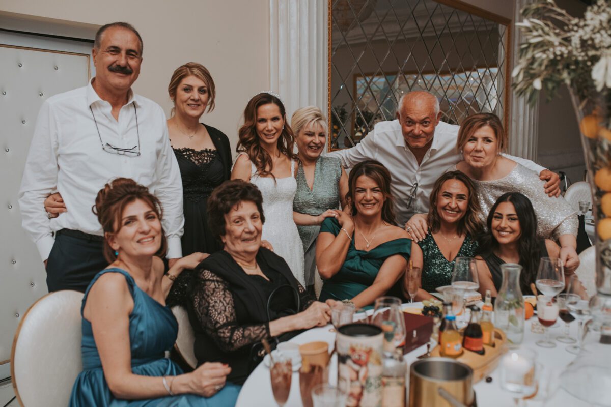 Eine Gruppe von Menschen versammelt sich bei einer Indoor-Veranstaltung, vermutlich einer standesamtlichen Trauung, um einen mit Essen und Getränken geschmückten Tisch. Einige stehen, andere sitzen, alle sind in formeller Kleidung gekleidet und haben meist fröhliche Ausdrücke. Im Hintergrund sind Blumendekorationen zu sehen und seitlich ist ein Kronleuchter zu sehen. - Elis Wedding - Hochzeitsfotograf Frankfurt