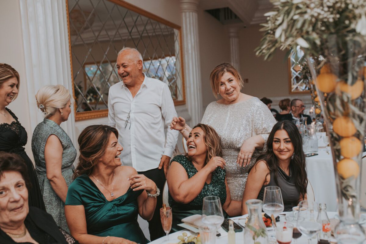 Eine Gruppe von Menschen versammelt sich bei einer formellen Veranstaltung um einen Esstisch mit verschiedenen Getränken und Gerichten. Vier Frauen, elegant gekleidet, sitzen und lächeln. Ein Mann in einem weißen Hemd und eine Frau in einem schimmernden Kleid stehen hinter ihnen und unterhalten sich angeregt – perfekte Motive für jeden Hochzeitsfotografen. - Elis Wedding - Hochzeitsfotograf Frankfurt
