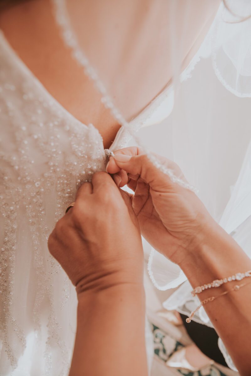 Nahaufnahme einer Person, die den Rücken des weißen, verzierten Hochzeitskleides einer Braut schließt. Das Kleid der Braut ist mit aufwendigen Perlenarbeiten und zartem Stoff verziert. Die mit Armbändern geschmückten Hände der Person konzentrieren sich während eines Fotoshootings eines Hochzeitsfotografen darauf, den Verschluss oder die Knöpfe des Kleides zu schließen. - Elis Wedding - Hochzeitsfotograf Frankfurt