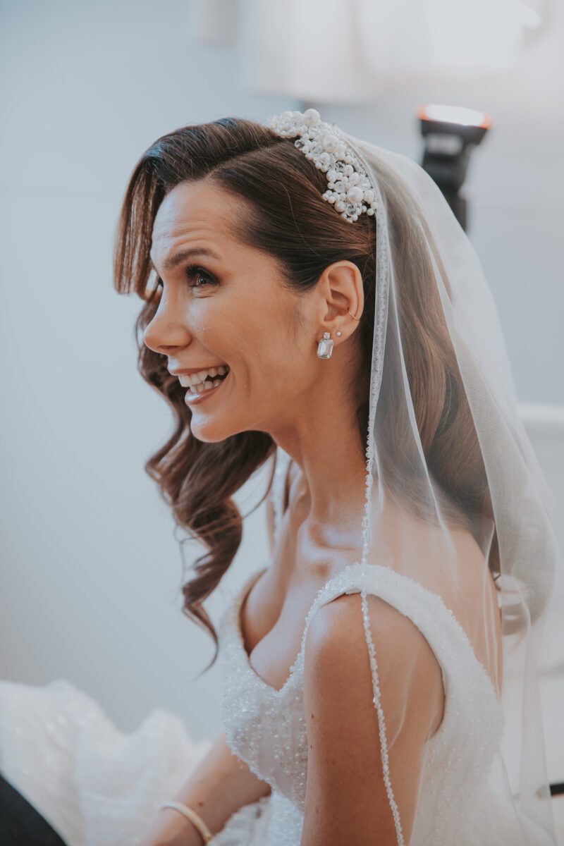 Eine Braut mit langen braunen, in lockeren Wellen gestylten Haaren lächelt strahlend. Sie trägt eine funkelnde Tiara und einen transparenten Schleier mit aufwendigen Details. Während des Fotoshootings ist ihr weißes Brautkleid mit zarten Perlen verziert. Perlenohrringe ergänzen ihren Look. Der Hintergrund ist sanft fokussiert und lenkt die Aufmerksamkeit auf ihren freudigen Gesichtsausdruck. - Elis Wedding - Hochzeitsfotograf Frankfurt