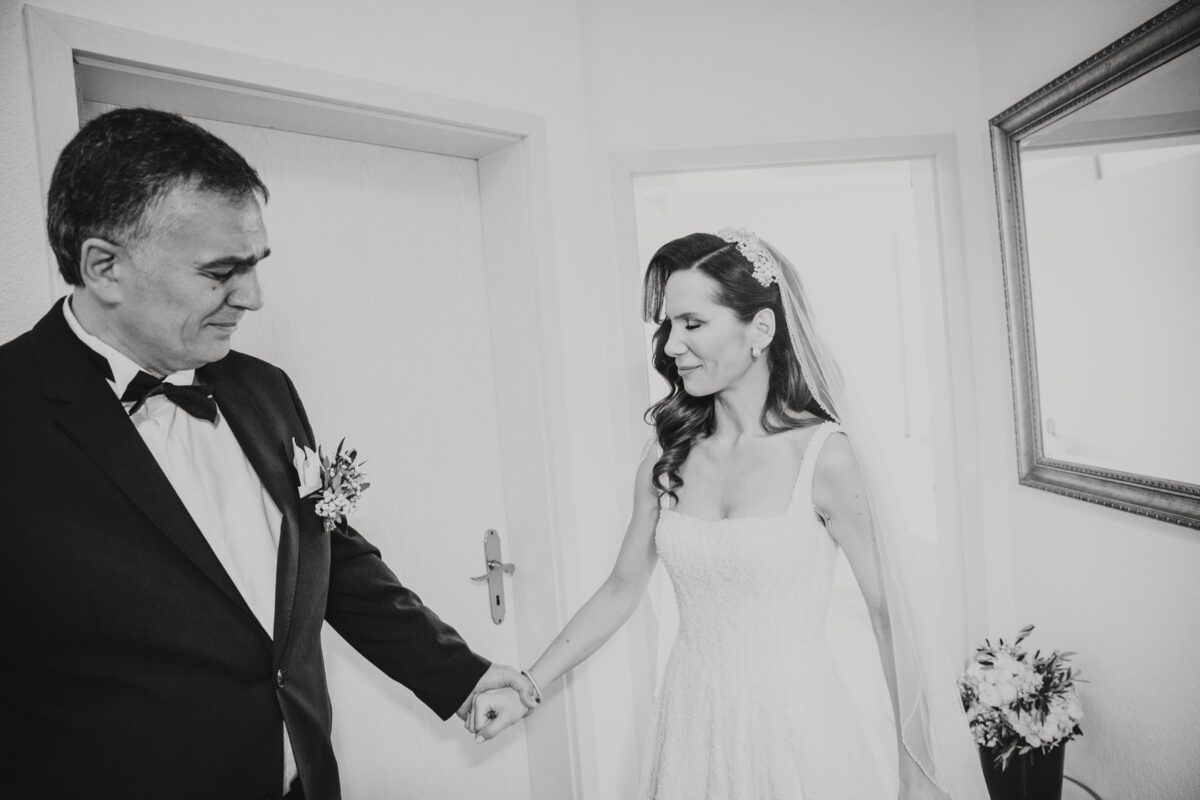 Eine Schwarz-Weiß-Fotografie zeigt eine Braut und einen Mann, die drinnen stehen. Die Braut in einem weißen Kleid mit einem Blumenstrauß in der Hand wirkt emotional, als sie die Hand des Mannes im dunklen Anzug mit einer Ansteckblume hält. Sie stehen in der Nähe einer Tür, in der ein Spiegel und ein Blumenarrangement zu sehen sind, vielleicht während ihrer standesamtlichen Trauung. - Elis Wedding - Hochzeitsfotograf Frankfurt