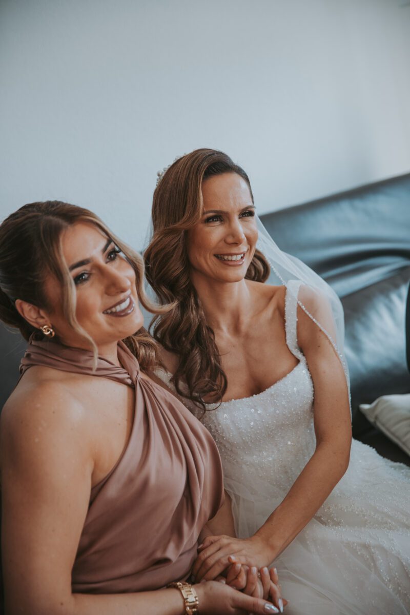 Zwei Frauen sitzen lächelnd und händchenhaltend auf einem dunklen Sofa. Eine trägt ein ärmelloses, pfirsichfarbenes Kleid mit drapiertem Ausschnitt und Goldschmuck, während die andere ein perlenbesetztes weißes Brautkleid mit Schleier trägt. Aufgenommen von ihrem Hochzeitsfotografen während der standesamtlichen Trauung, haben beide langes, welliges Haar und blicken aus dem Kamerablickwinkel. - Elis Wedding - Hochzeitsfotograf Frankfurt