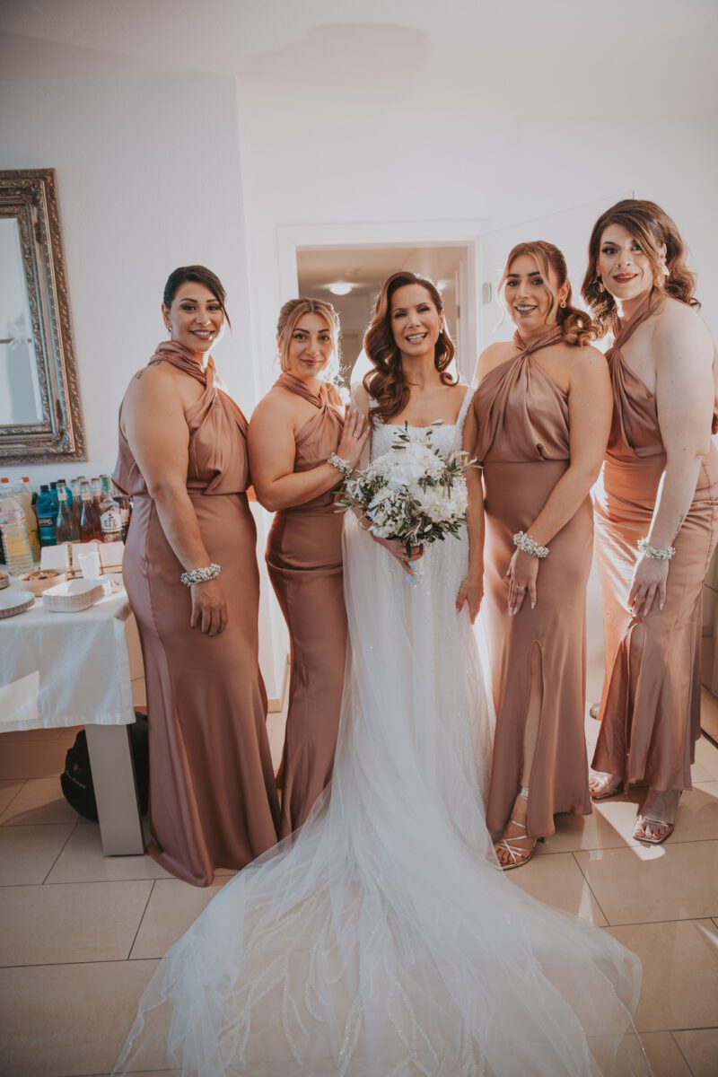 Eine Braut in einem weißen Brautkleid mit einem Blumenstrauß steht lächelnd neben vier Brautjungfern in passenden roségoldenen Satinkleidern. Sie posieren für professionelle Fotografie im Innenbereich vor einer Tür. Links daneben steht ein kleiner Tisch mit einer Auswahl an Getränken und persönlichen Gegenständen. Der Raum ist hell erleuchtet. - Elis Wedding - Hochzeitsfotograf Frankfurt