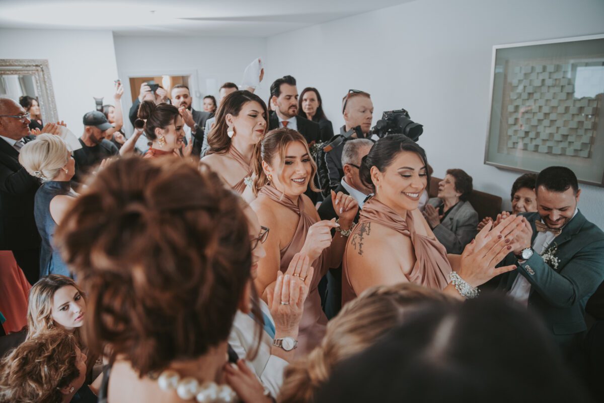 Ein überfüllter Raum voller feiernder Menschen. Mehrere Frauen in eleganten Kleidern und ein Mann im Anzug klatschen freudig. Die Atmosphäre ist lebhaft, perfekt für einen Hochzeitsfotografen, um den Moment festzuhalten. Der Raum verfügt über einen großen Spiegel auf der linken Seite, abstrakte Kunst an der rechten Wand und natürliches Licht fällt hinein. - Elis Wedding - Hochzeitsfotograf Frankfurt