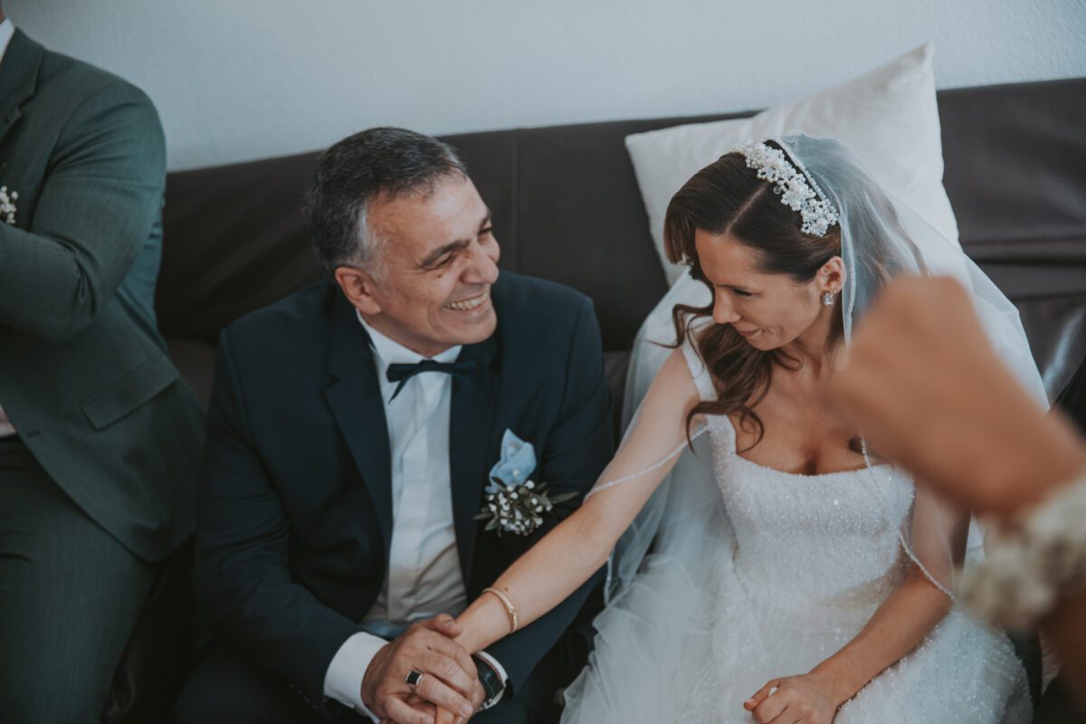 Eine Braut in einem weißen Brautkleid und Schleier sitzt neben einem Mann in einem dunklen Anzug mit Fliege. Sie halten Händchen und lächeln sich an. Die Braut trägt einen mit Juwelen besetzten Kopfschmuck und der Mann eine Ansteckblume. Aufgenommen während ihrer standesamtlichen Trauung, ist auf der rechten Seite des Bildes teilweise der Arm einer weiteren Person zu sehen. - Elis Wedding - Hochzeitsfotograf Frankfurt