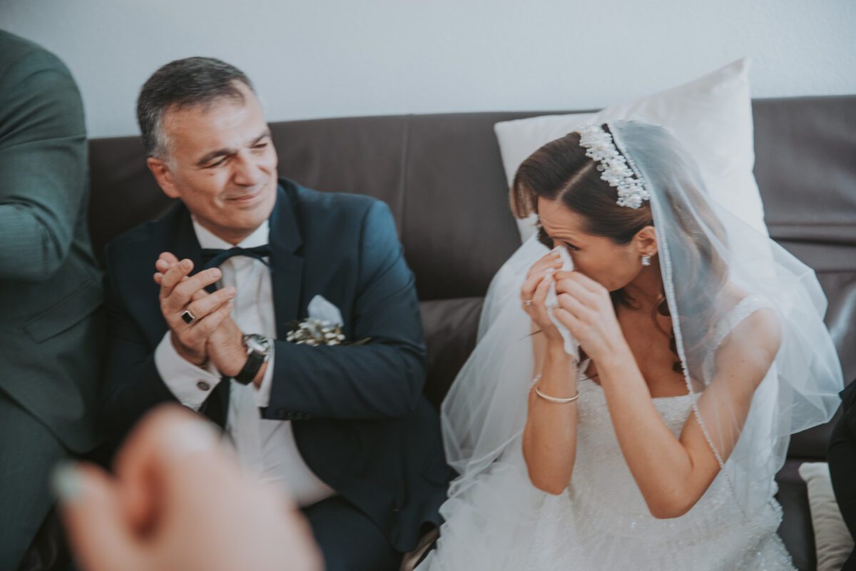 Eine Braut in weißem Brautkleid und Schleier sitzt auf einem Sofa im Standesamtlichen Trauung in Frankfurt und hält sich ein Taschentuch vors Gesicht, während sie weint. Neben ihr klatscht ein Mann in dunklem Anzug und Fliege und lächelt sie an. Sie sind umgeben von einem Innenraum mit hellen Wänden. - Elis Wedding - Hochzeitsfotograf Frankfurt