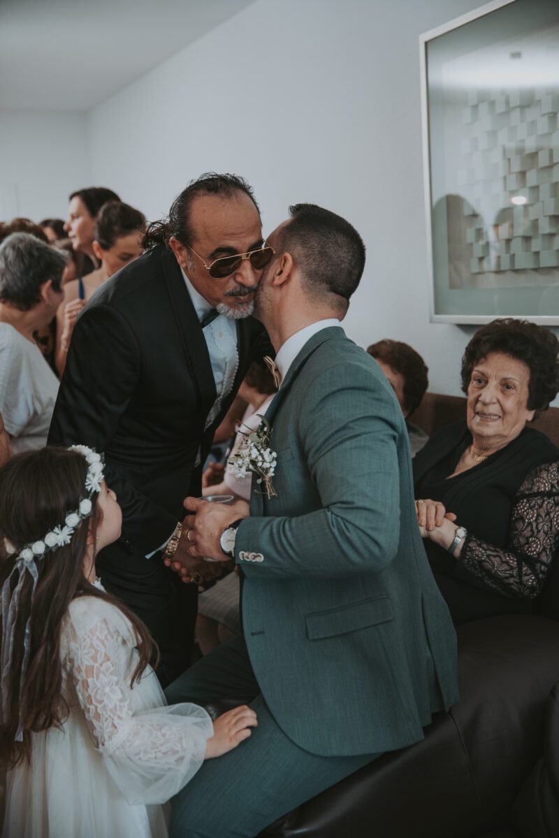Ein Mann im Anzug mit einer Blumenboutonnière sitzt da, schüttelt einem älteren Mann im schwarzen Anzug die Hand und bekommt einen Kuss auf die Wange. In der Nähe steht während eines Fotoshootings ein junges Mädchen in einem weißen Kleid mit einer Blumenkrone. Eine ältere Frau in einem schwarzen Kleid mit Spitzenärmeln beobachtet die Szene, während sie auf einem Sofa sitzt. - Elis Wedding - Hochzeitsfotograf Frankfurt