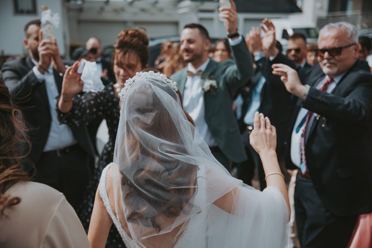 Eine Braut in weißem Kleid und Schleier, die den Blick abwendet, ist von formell gekleideten Menschen umgeben. Sie stehen im Freien, lächeln und heben feierlich die Hände. Im Hintergrund sind Gebäude und ein teilweise klarer Himmel zu sehen. Die Stimmung ist festlich und fröhlich, perfekt eingefangen vom Hochzeitsfotografen. - Elis Wedding - Hochzeitsfotograf Frankfurt
